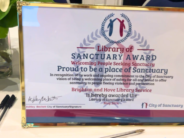 The image shows the Library of Sanctuary award in a golden frame propped up on a table.