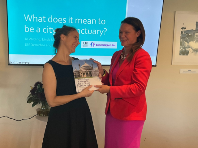 The image shows a smiling Council Leader Bella Sankey wit Sussex University researcher Jo Wilding, holding the 'What does it mean to be a City of Sanctuary?' report.