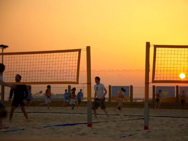 As the sun sets in an orange/pink sky two beach volleyball nets can be seen on a beach. One the left court two players on the far side of the net are poised ready for their next strike.