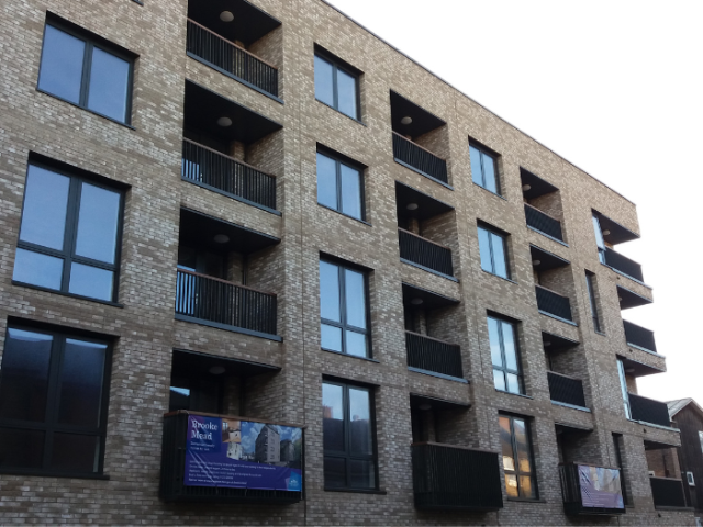 A 4 storey new block of flats with balconies on the far rigth hand corner. Each storey has 3 sets of square windown units separated by square balcony sections.