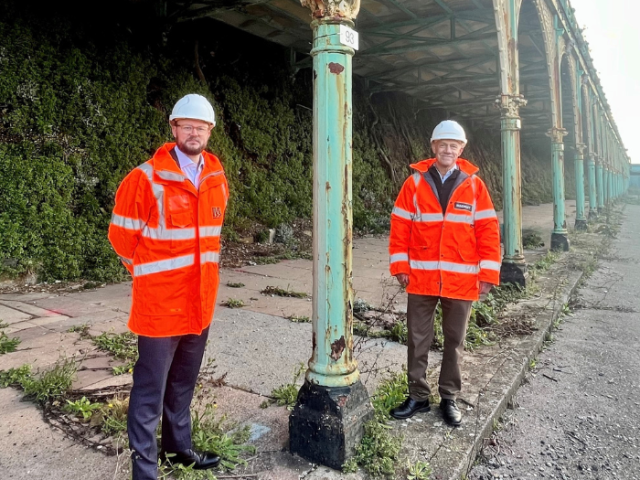 Councillor Jacob Taylor adn MAckley MD Ben Hamer, oth dressed in orange high viz jackets and white helmets stand by a light lbue cast iron pillar of Madeira terrace. 