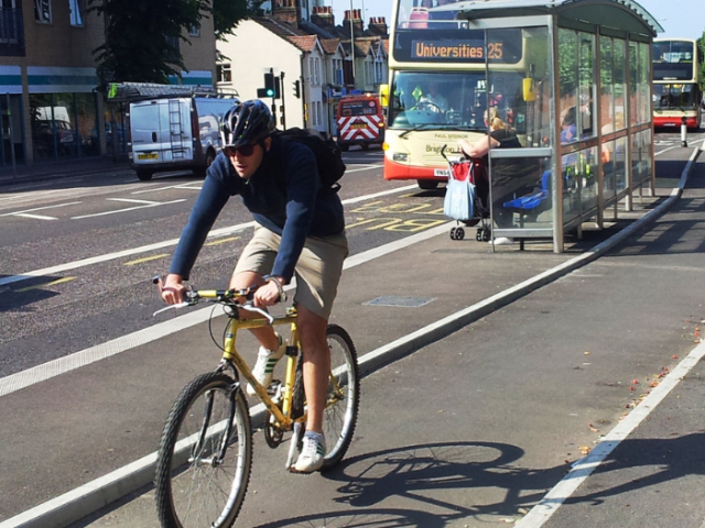 A cyclyst in a bluye helmet and top is riding a yellow-framed bike ibn a cycle lane toward the camera. Behind him a bus is sat at a bus stop with another behind it.