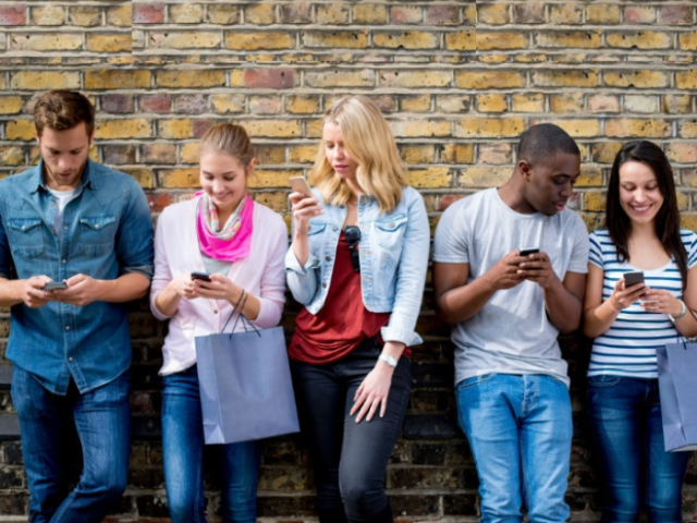 A group ofyoung people have their backs to a brick wall and are looking at their mobile phones and chatting to those next to them. 