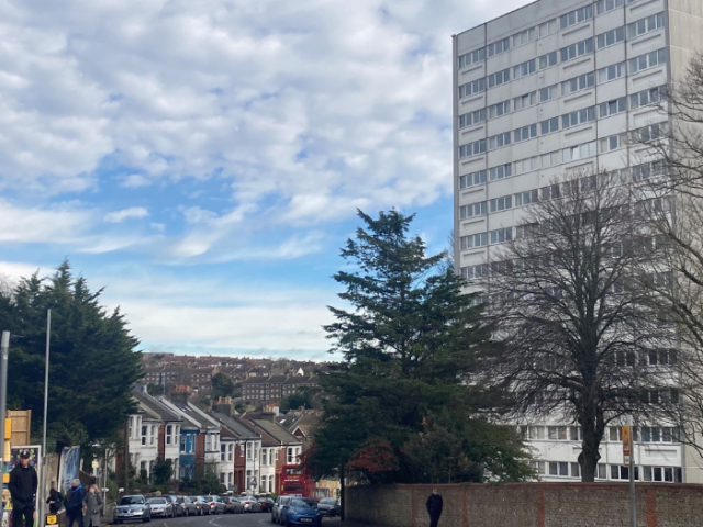 A view across Brighton shows a multi-story block on the right hand side of a street, with a terrace of 2 story houses opposite it. Seceral tress front the buildings on either side of the road.