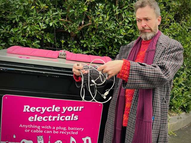 Councillor Rowkins untangles some cables before putting them in the small electricals recycling bin. Cllr Rowkins is standing next to the bin which has a bright pink sign with the words 'Recycle Your Electricals' on it.