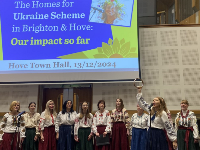 The image shows the Ukrainian Voices Choir singing in Hove Town Hall.