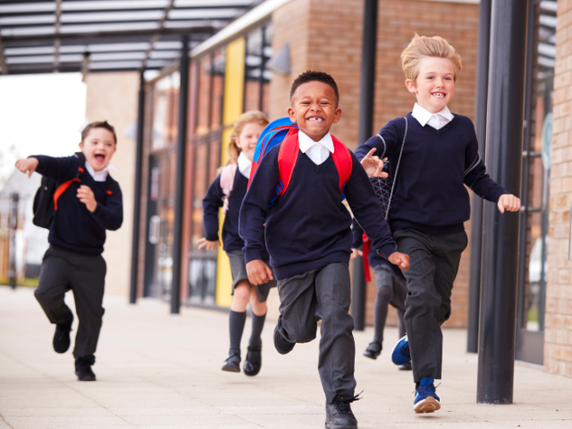 A group of children wearing school uniforms and rucksacks running in front of a building