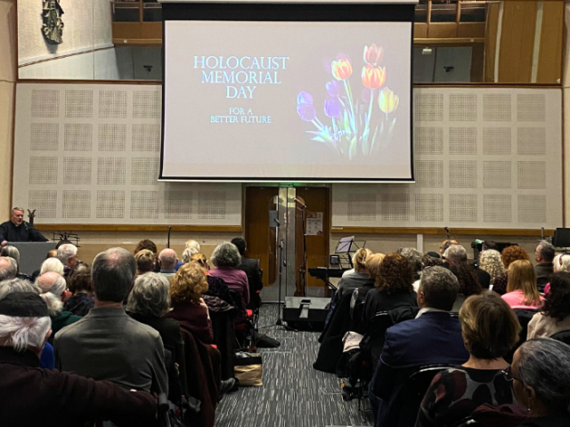 The image shows a crowd of people sat in chairs, looking at a screen. The projection on the screen reads: Holocaust Memorial Day. For a better future.