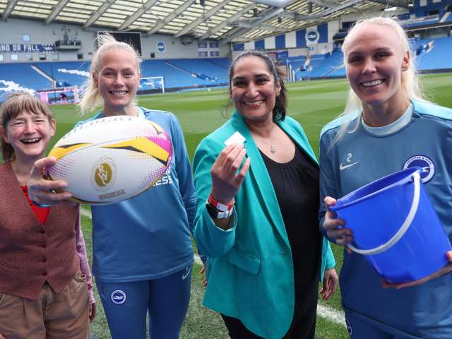 Councillor Mitchie Alexander, Guro Bergsvand, Manjinder Nagra and Maria Thorisdottir at Brighton & Hove Albion's stadium. Guro is holding a rugby ball and Manjinder has one of the winning tickets. 