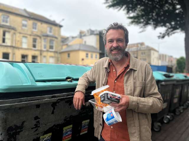 Councillor Tim Rowkins holding some pots, tubs and trays next to a blue-lidded communal recycling bin.