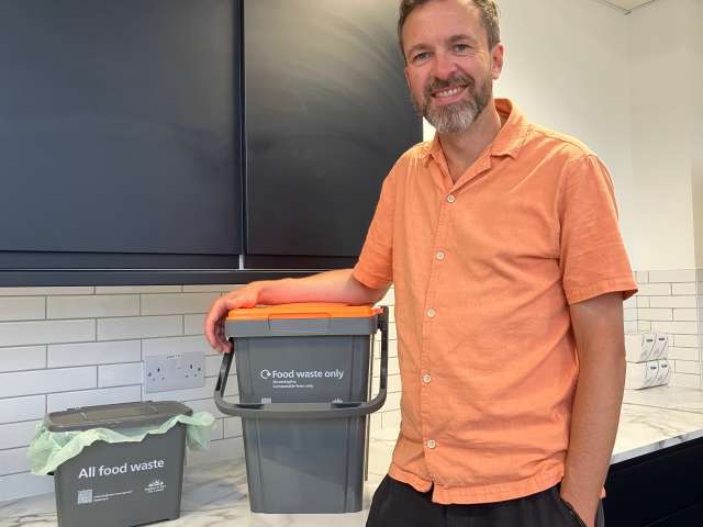 Councillor Tim Rowkins standing in a kitchen next to cupboards wearing a short-sleeved orange shirt, with a small grey kitchen caddy and larger outdoor caddy with orange lid on the kitchen worktop