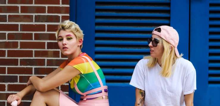 Two women sitting by a brick wall. One is wearing a rainbow striped top and a pink skint, the other is wearing a pink baseball cap backwards and a white t shirt