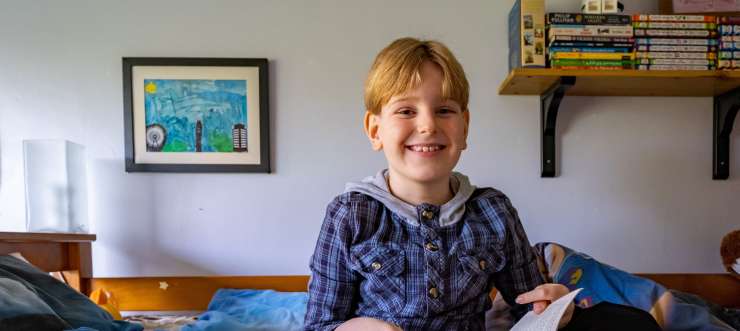 Young boy sitting on a bed in his room smiling and holding an open book