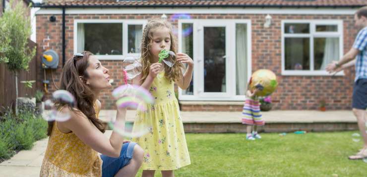 Adult woman blowing bubbles with young child whilst adult man plays ball with a toddler. They are all in a back garden.