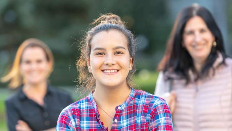 Teenager smiling into the camera. 2 adults out of focus in the background