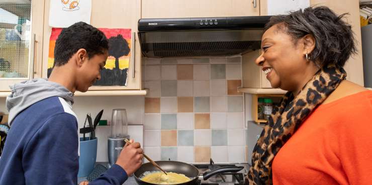 An adult and teenager in a kitchen. The teenager is cooking an omelette. The adult is smiling whilst looking at the teenager.