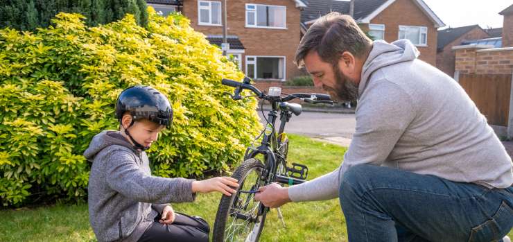 Adult helping young child inflate the tyres on his bike. The young child is wearing a bike helmet. The adult and child are sitting on the grass in a front garden. There are other housing in the background 