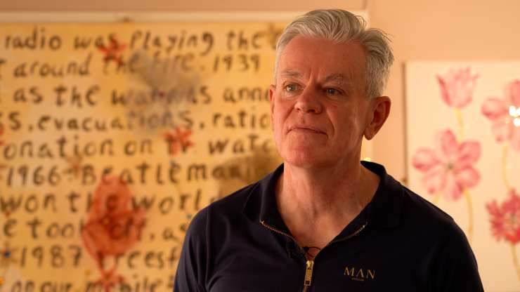 Close up of a person facing the camera, there is some picture on the wall behind with flowers and words on.