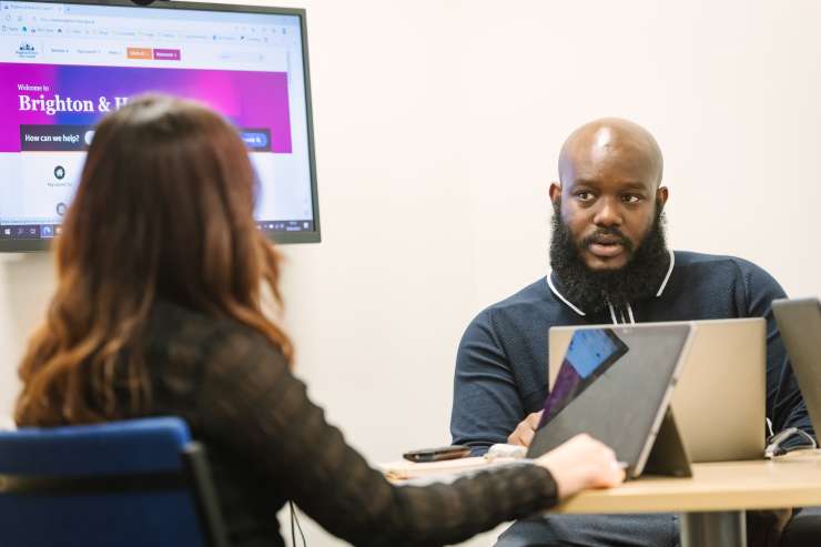 2 people sitting in an office, facing each other. There is a screen in the background showing the Brighton & Hove City Council logo.