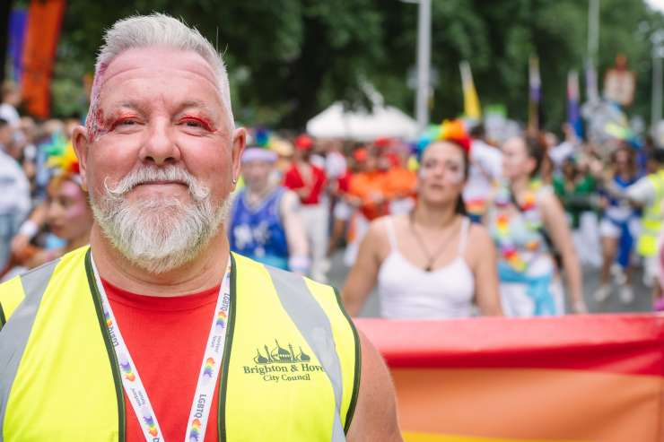 In the foreground is a Brighton & Hove City Council worker wearing a yellow high-vis vest, they have glitter on their face and colourful eye shadow. They are at the Pride celebration and in the background are party goers in a group behind a barrier. 