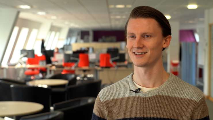 A person is standing in an office smiling, in the background there are banks of office desks with red office chairs.