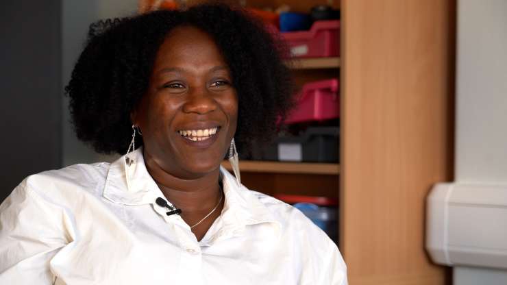 Close up of a person sitting in an office, they are facing the camera and smiling, there is a bookshelf in the background.