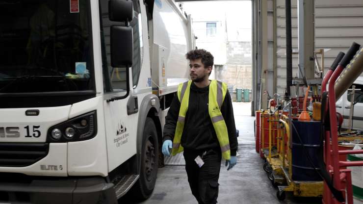 A mechanic is in a garage wearing a yellow high-vis vest, they are walking towards the camera, to their right is a large white Brighton & Hove City Council lorry, to their left there are barrels and trolleys.