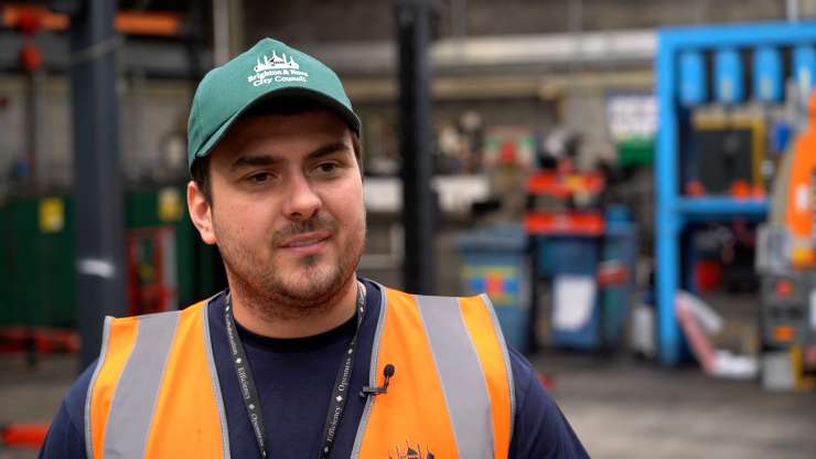 A mechanic wearing an orange high-vis vest and a green Brighton & Hove City Council cap is facing the camera smiling. They are in the entrance of a garage with shelves of tools behind them. 