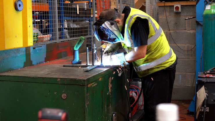 A mechanic is soldering at a workbench in a workshop, they are wearing a large protective mask and there are spark flying up towards them.