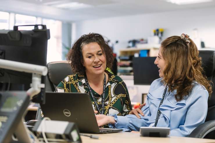 2 people sitting at an office desk talking and smiling at each other. There is a laptop open on the desk in front of them.