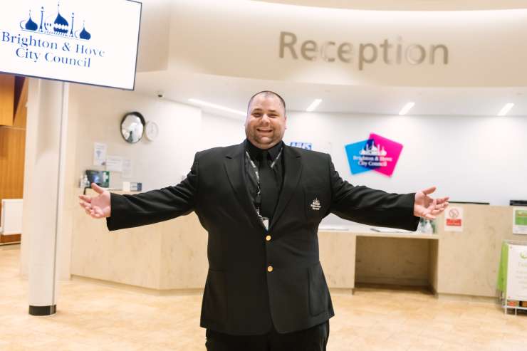 A Brighton & Hove City Council security guard is standing in Hove Town Hall reception, they are facing the camera with a big smile on their face and their arms stretched out wide. In the background is the reception desk and the Brighton & Hove City Council logo on the wall. 