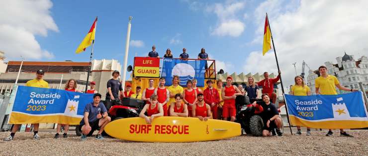 The Brighton & Hove lifeguard team are assembled on the beach to pose for a group photo. The sky behind them is blue with a few fluffy clouds. 